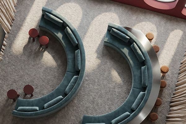 Curved teal couches face each other on a light carpet with round red side tables. Sunlight casts patterns, creating a calm, inviting atmosphere.