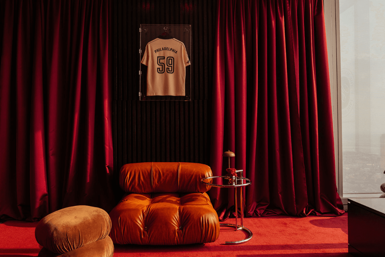Cozy room with red velvet curtains, a framed sports jersey on a black wall, a tan leather chair, a matching footrest, and a small side table.