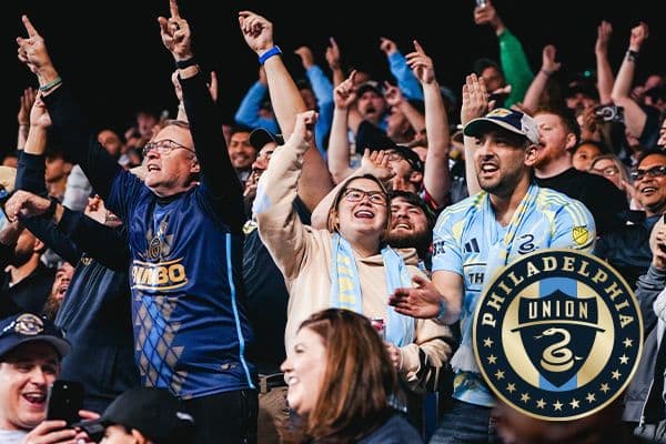 A group of enthusiastic Philadelphia Union soccer fans cheer in the stands, wearing team jerseys and scarves, creating an energetic and joyful atmosphere.